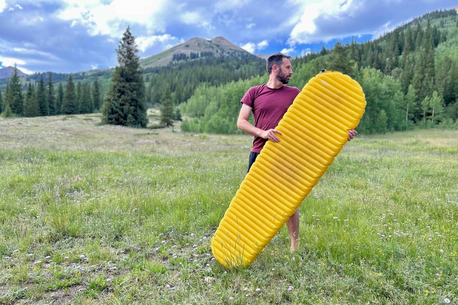 A man stands outdoors, holding a large yellow sleeping pad, with forest and mountains in the background.