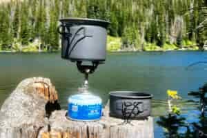 A backpacking stove with a pot on top sits on a stump in front of a mountain lake on a sunny day.