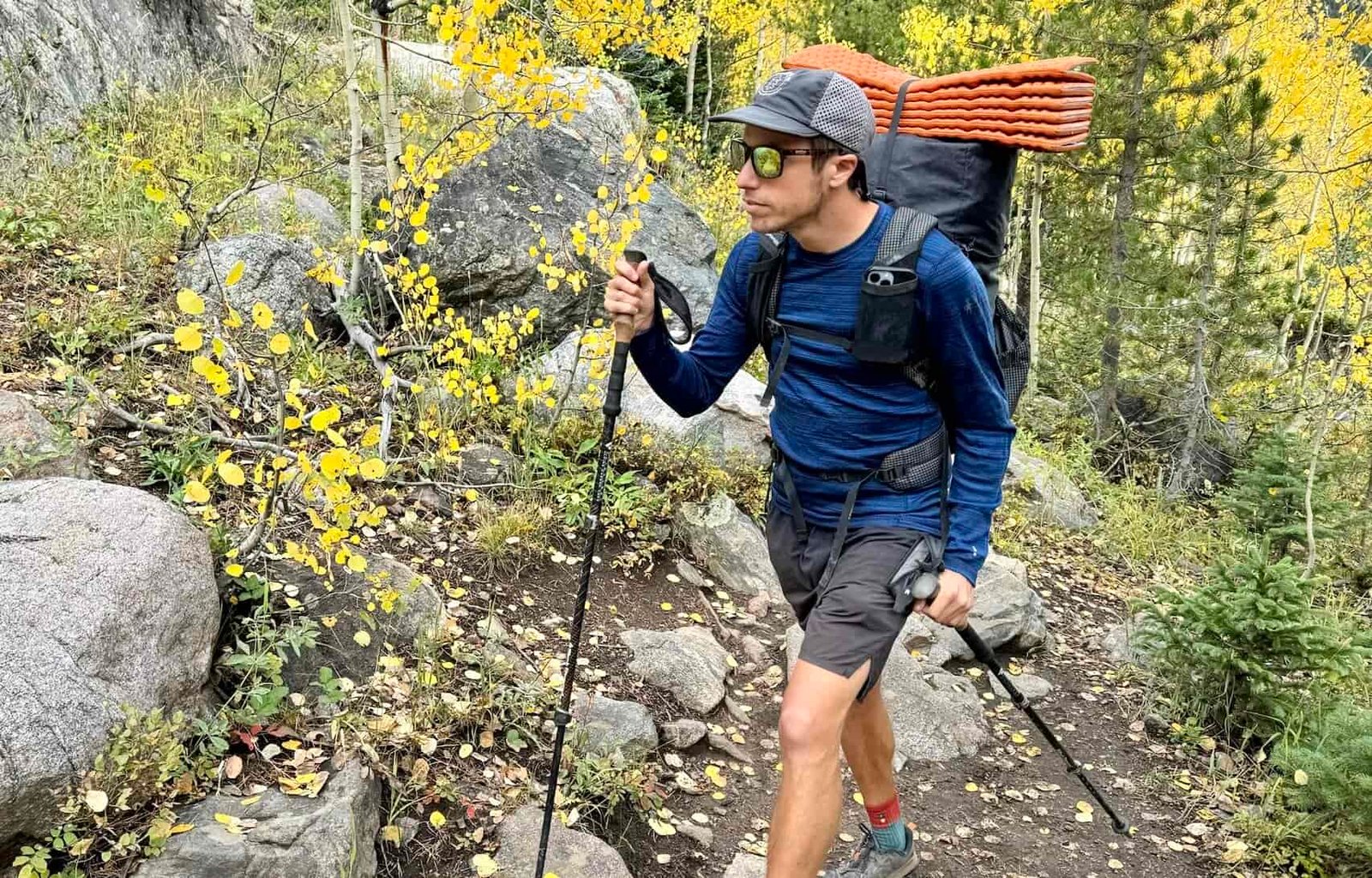 A hiker walks through the woods with trekking poles and a pack while wearing the Smartwool Classic Thermal base layer. Trees and rocks are in the background.