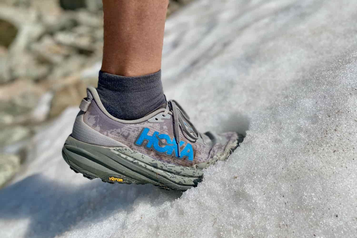 A hiker wearing the HOKA Speedgoat 6 trail running shoes while stepping in snow on a hike up a mountain.