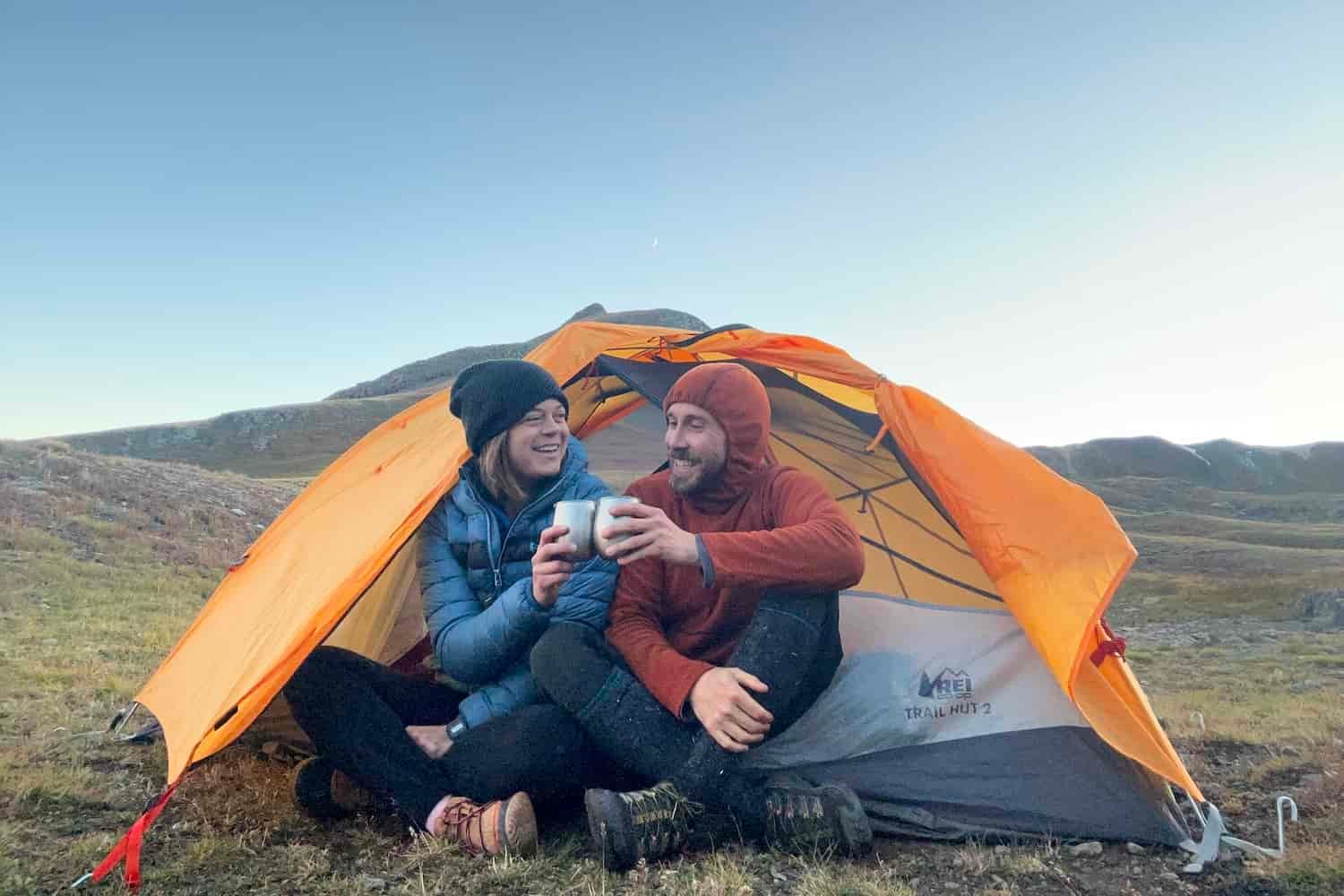 A man and a woman sit in the door of a tent cheersing two metal cups as the sun sets over the. mountains behind them.