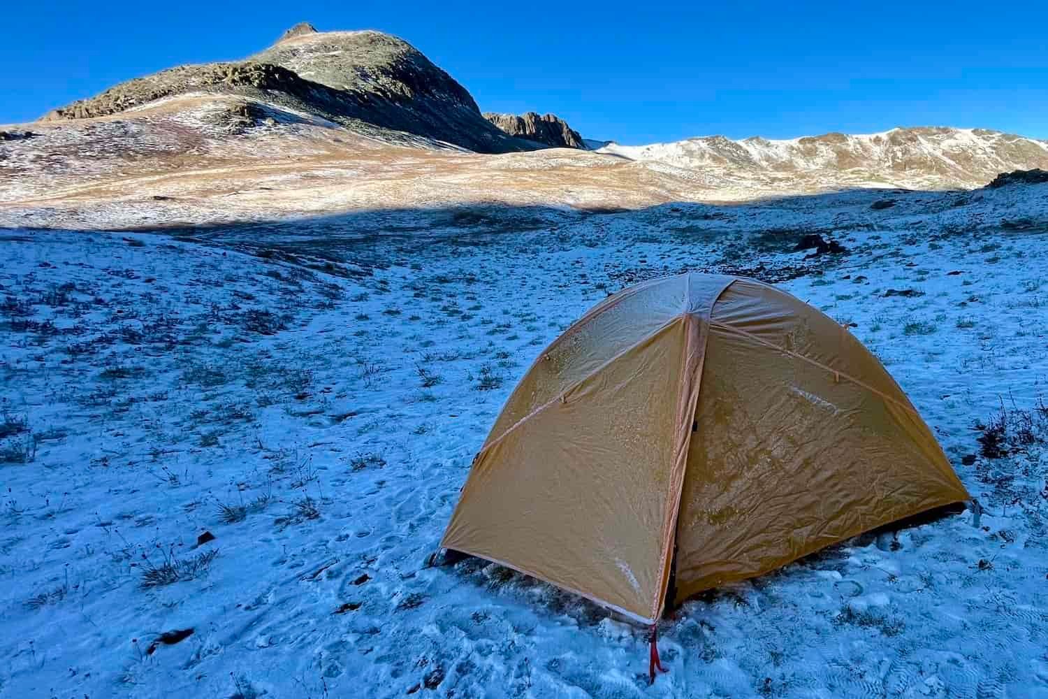 A tent with a fly on sits in a snowy field below a mountain in shadow under a bright blue sky.