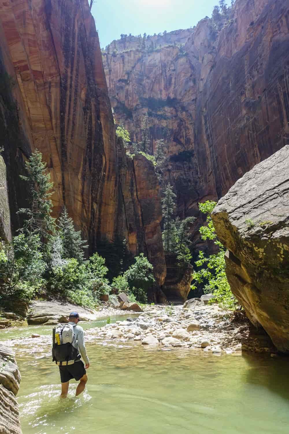 a backpacker walking in the sun through ankle deep water in a wider section of the narrows with redrock walls and boulders on all sides of the washout