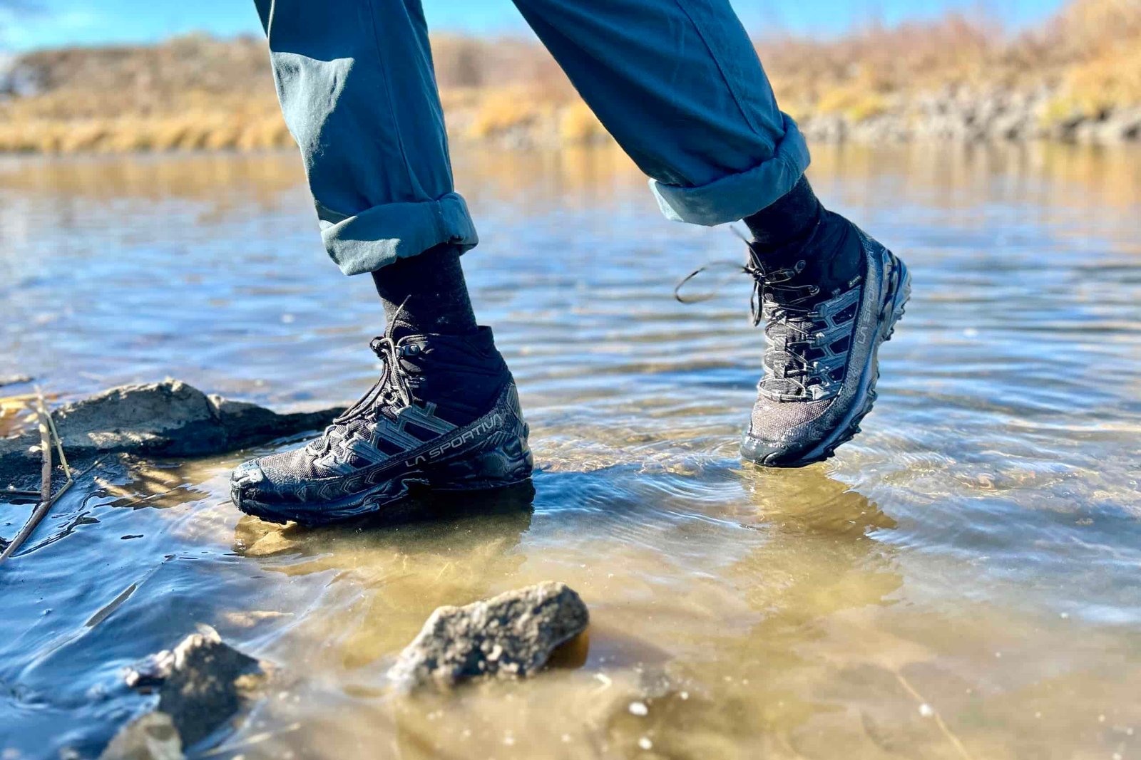 Close-up image from the knees down of a person walking in hiking boots through some water.