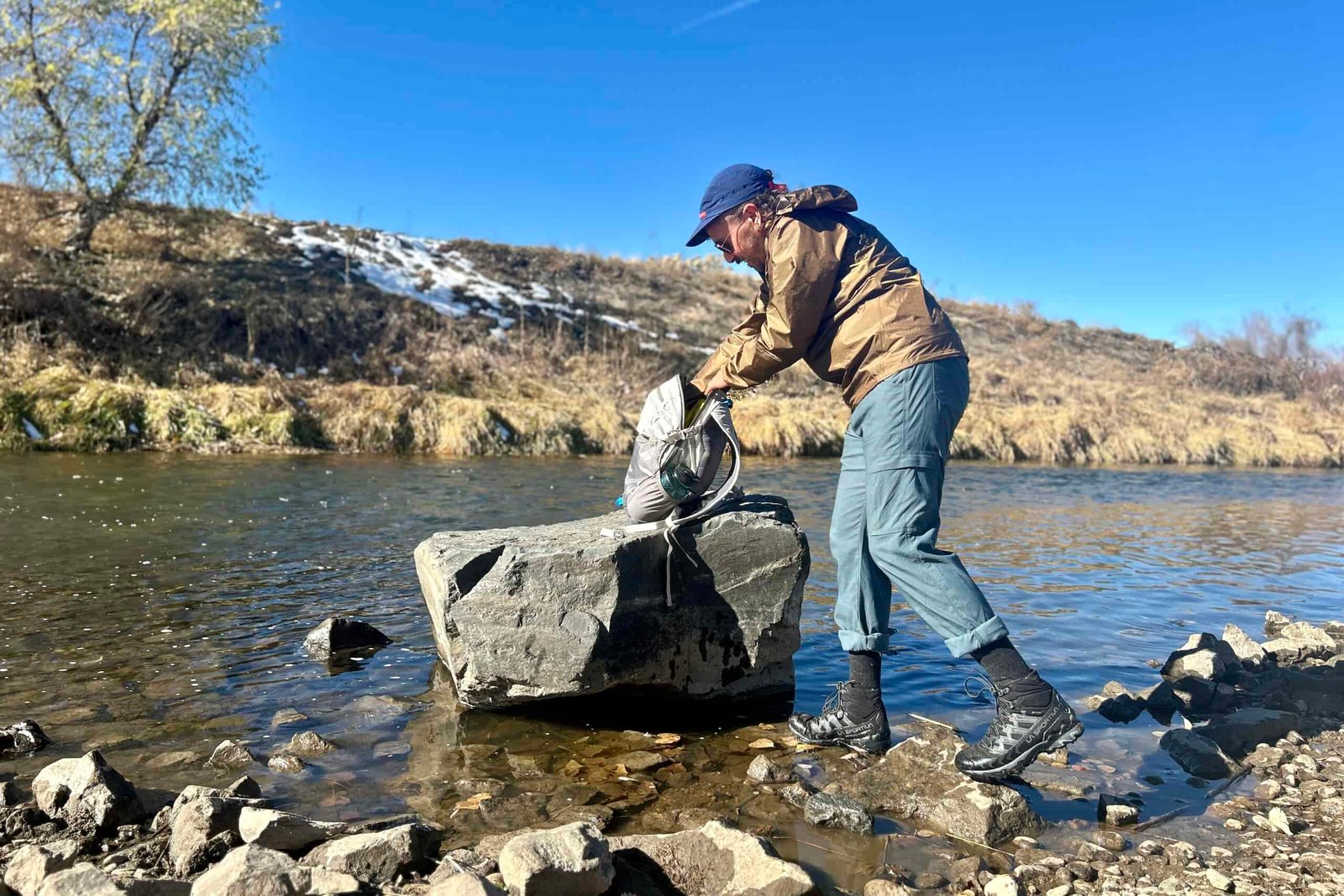 A man digs through his backpack along a river in a wilderness area in fall.