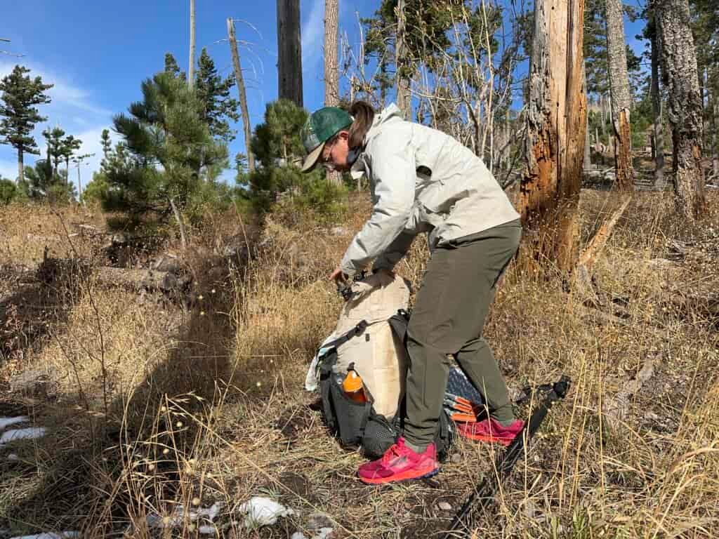 A hiker is packing up a backpack while wearing the Happy Hike pants in the woods.