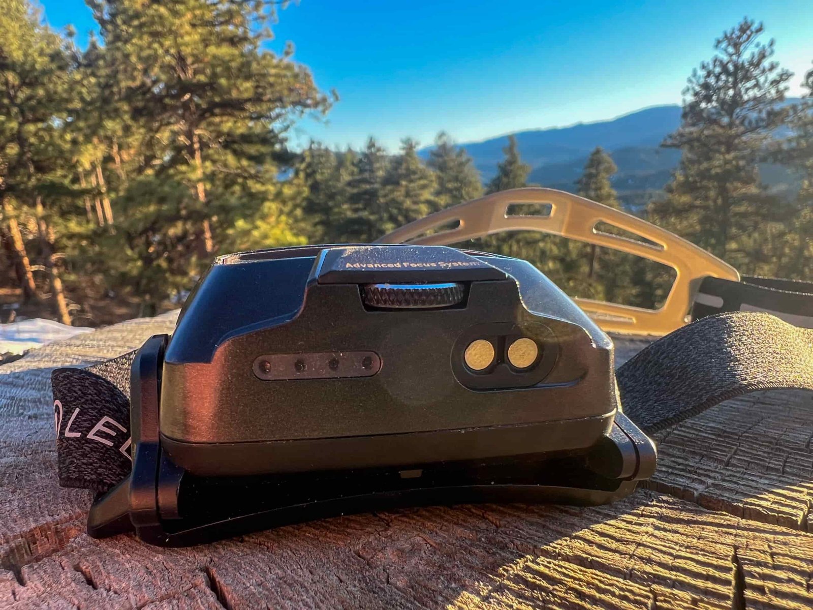 The image shows a close-up of the back of the Ledlenser HF6R Signature headlamp, highlighting the charging connection port and power indicator lights. The headlamp is placed on a wooden surface with a scenic background of trees and mountains. The headlamp's strap is visible on the left side, with a portion of the adjustable headband in the frame.