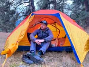 A camper sits cross-legged in the entrance of a brightly colored tent, showcasing its low headroom and cozy interior. The vibrant orange, yellow, and blue tent is pitched in a wooded area, with a pair of hiking boots placed on the grassy ground outside.