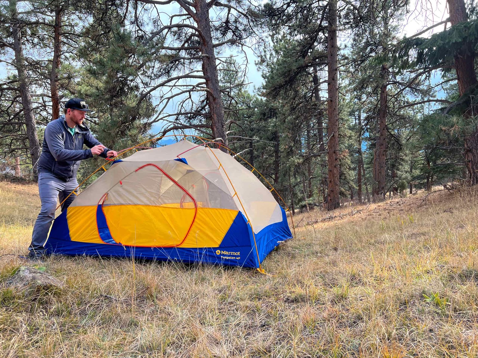 A camper setting up the Marmot Tungsten 4p tent in a wooded campsite, showcasing the tent's easy assembly with its color-coded pole system and spacious design.