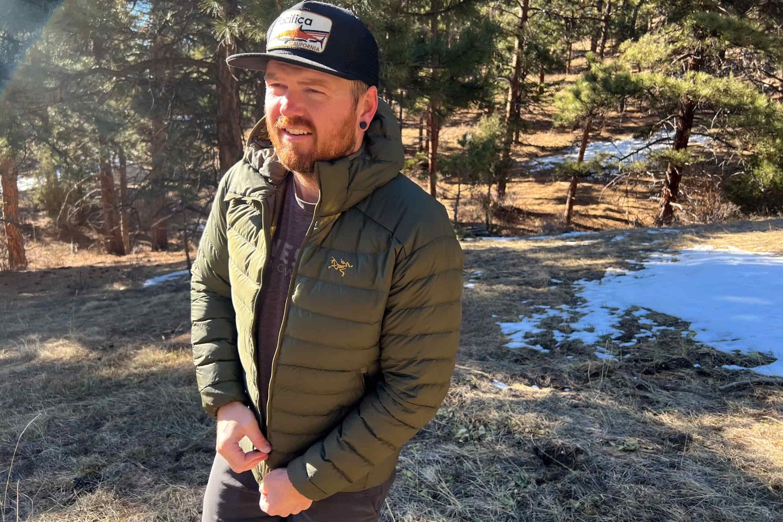 A man walking in the forest. He's zipping his jacket and looking past the camera. There are small snow patches in the background.