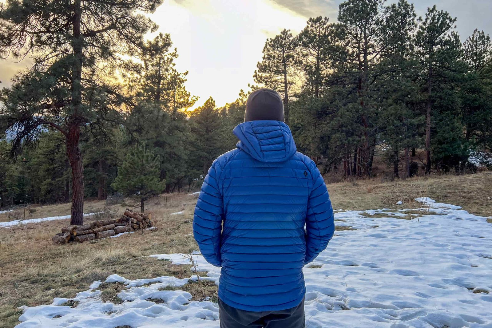 A person standing in a snowy field at sunset, showing the back view of the Black Diamond Approach Down Hoody with its hood up.