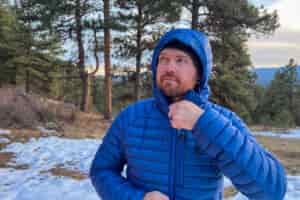 A person adjusting the zipper on the Black Diamond Approach Down Hoody, with the hood up, standing outdoors in a snowy forest setting.