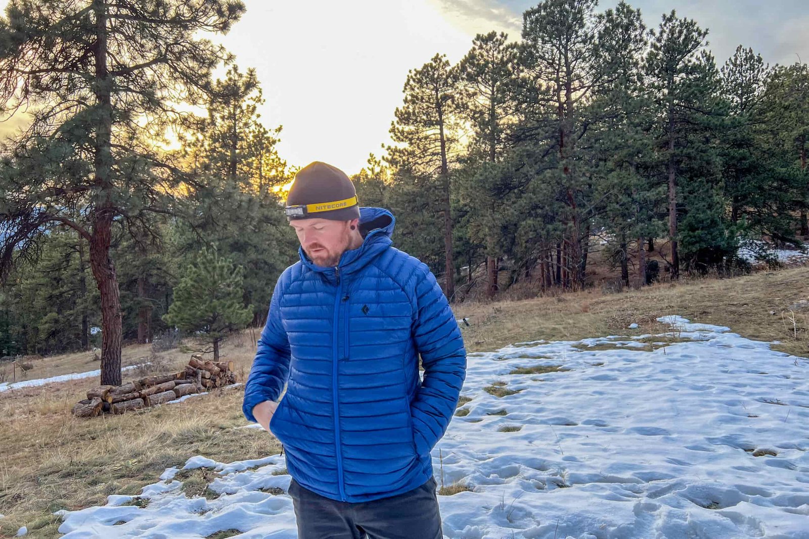 A person wearing the Black Diamond Approach Down Hoody, standing on a snow-dusted trail at sunset with pine trees and hills in the background.
