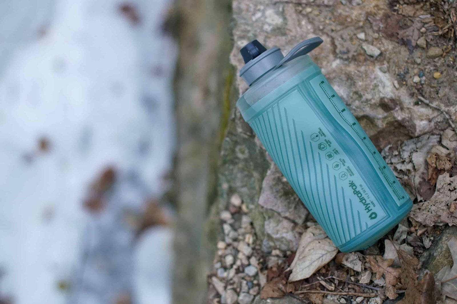 The Hydrapak Flux water bottle sits on gravel and leaves with snow in the background.
