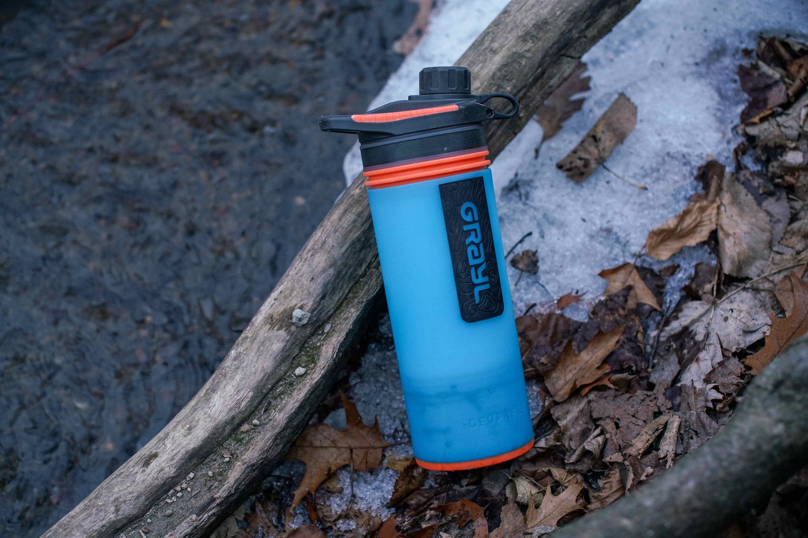 The Grayl GeoPress lays against a branch with a stream, snow, and leaves beneath it.