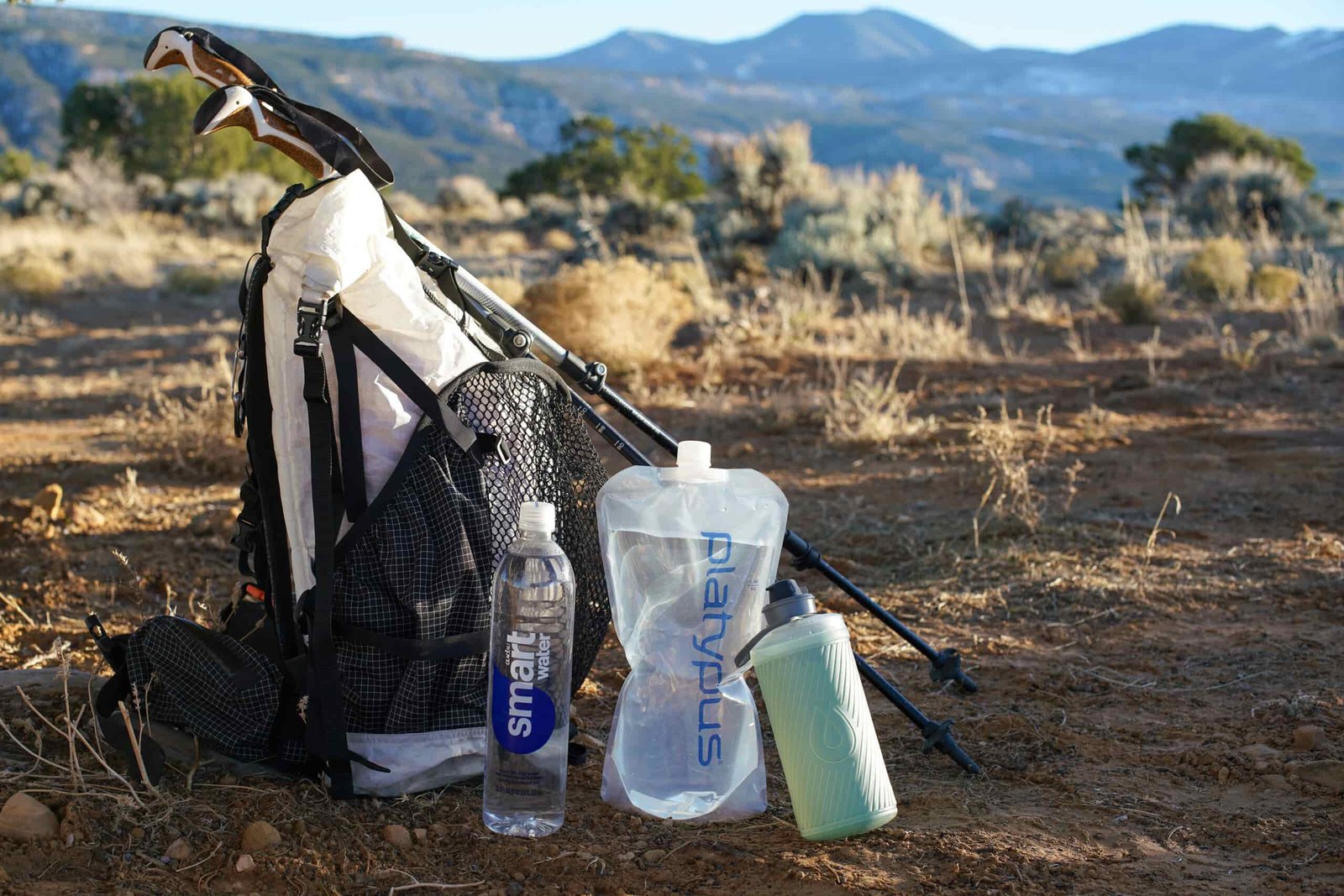 A bottle of Glaceau Smartwater, the Platypus Platy Bottle, and the Hydrapak Flux sit on the ground beside a backpack and trekking poles with mountains in the background.