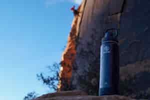 The Takeya Actives Insulated water bottle sits on a rock with a climber on a rock face behind the bottle.
