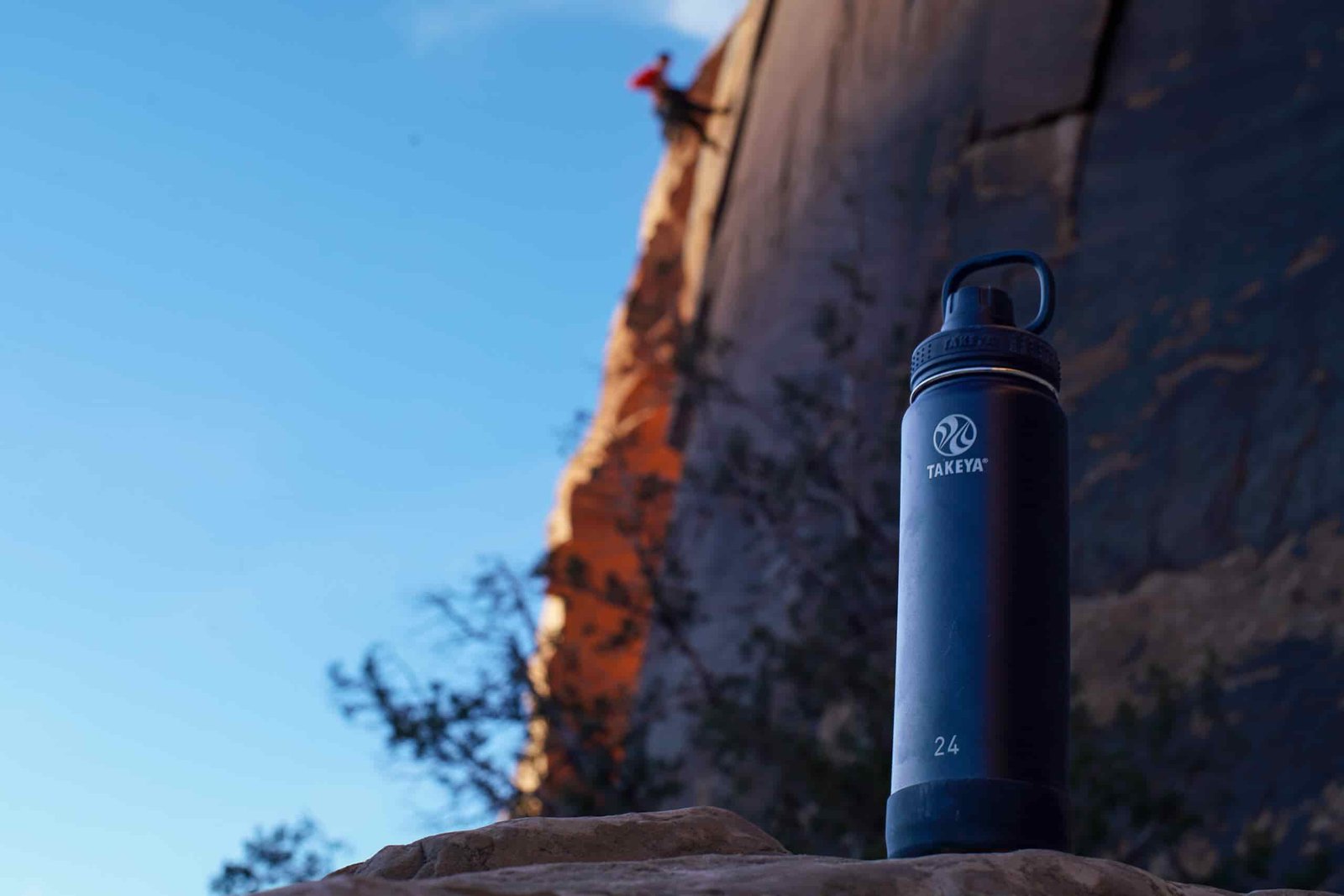 The Takeya Actives Insulated water bottle sits on a rock with a climber on a rock face behind the bottle.