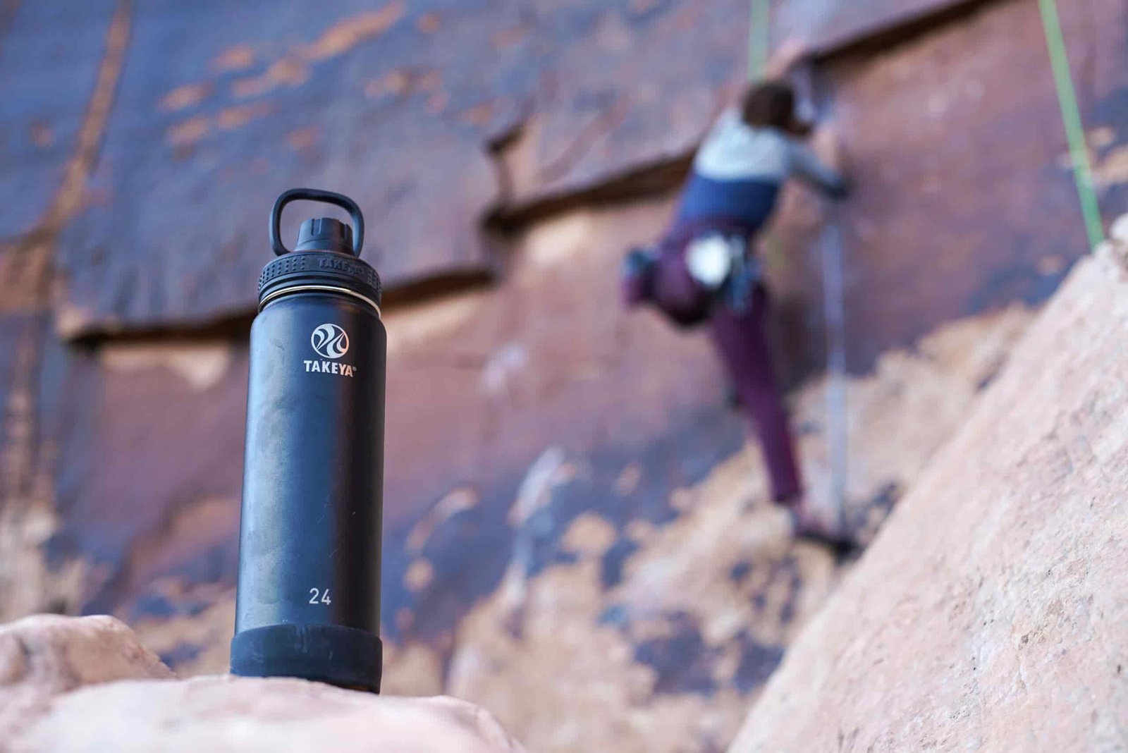 The Takeya Actives Insulated water bottle sits on a rock with a climber on a rock face behind the bottle.