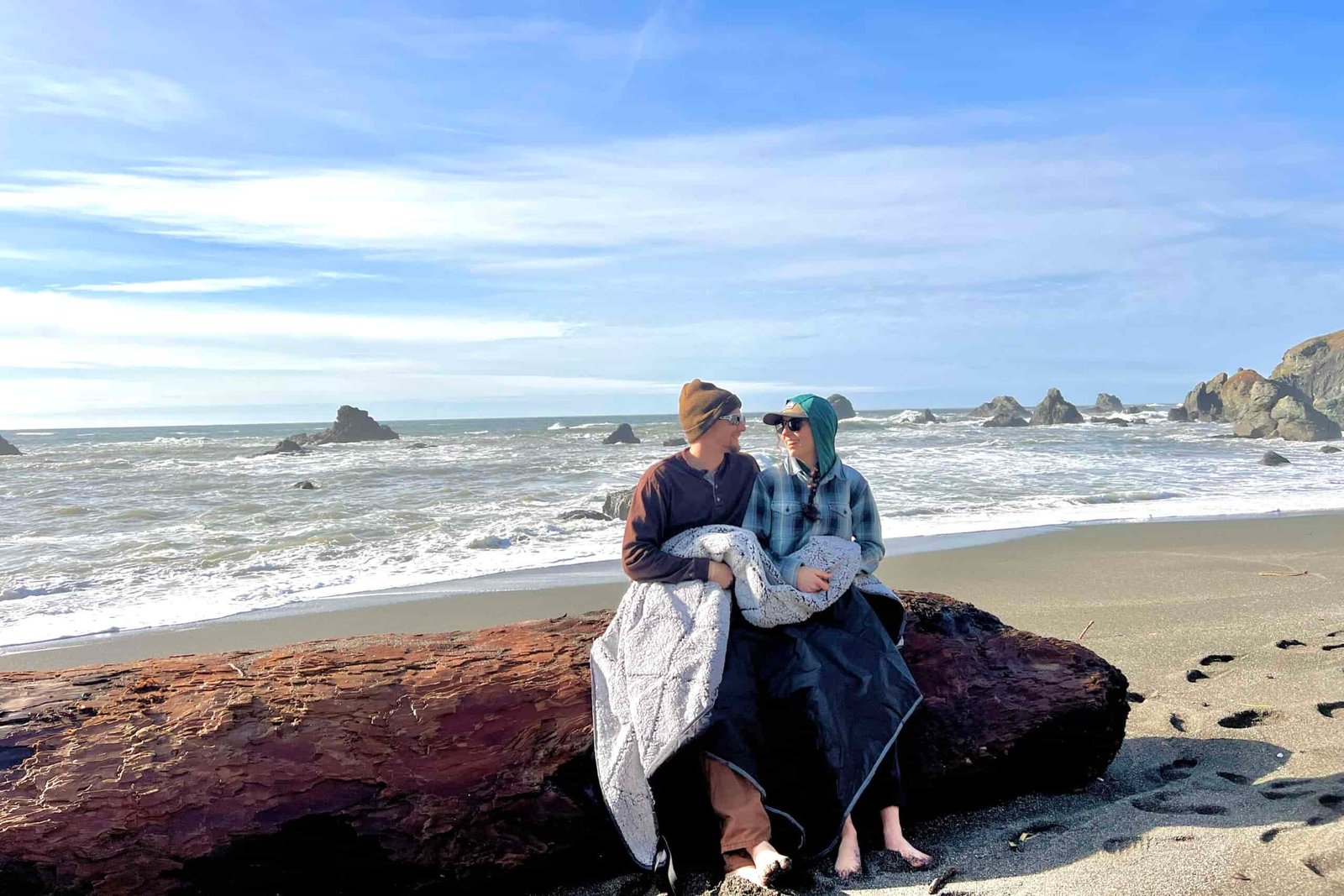 2 people sitting on a log at the beach, using the Oceas Sherpa Fleece Blanket as a throw blanket over their legs.