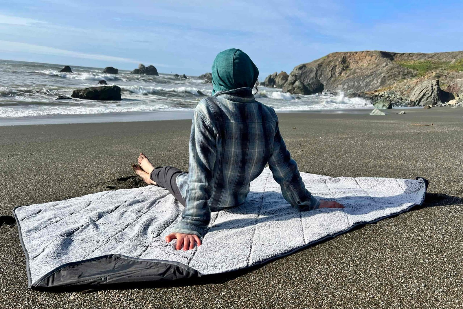 Person sits on the Oceas Sherpa Fleece Blanket at the beach, showing both the fleece top and the waterproof bottom layer.