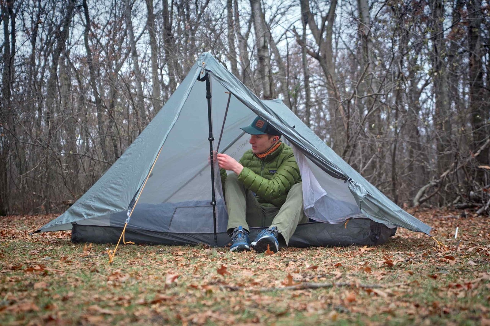 a man in a green coat puts his shoes on while sitting in a trekking pole tent