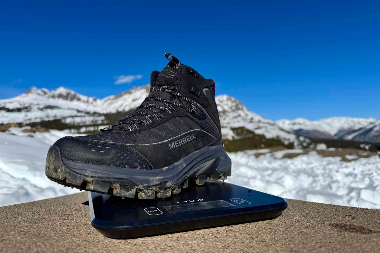 A close up of the Merrell boot sitting on a kitchen scale. The scale is sitting on a concrete wall in the mountains and there is snow covering the ground.
