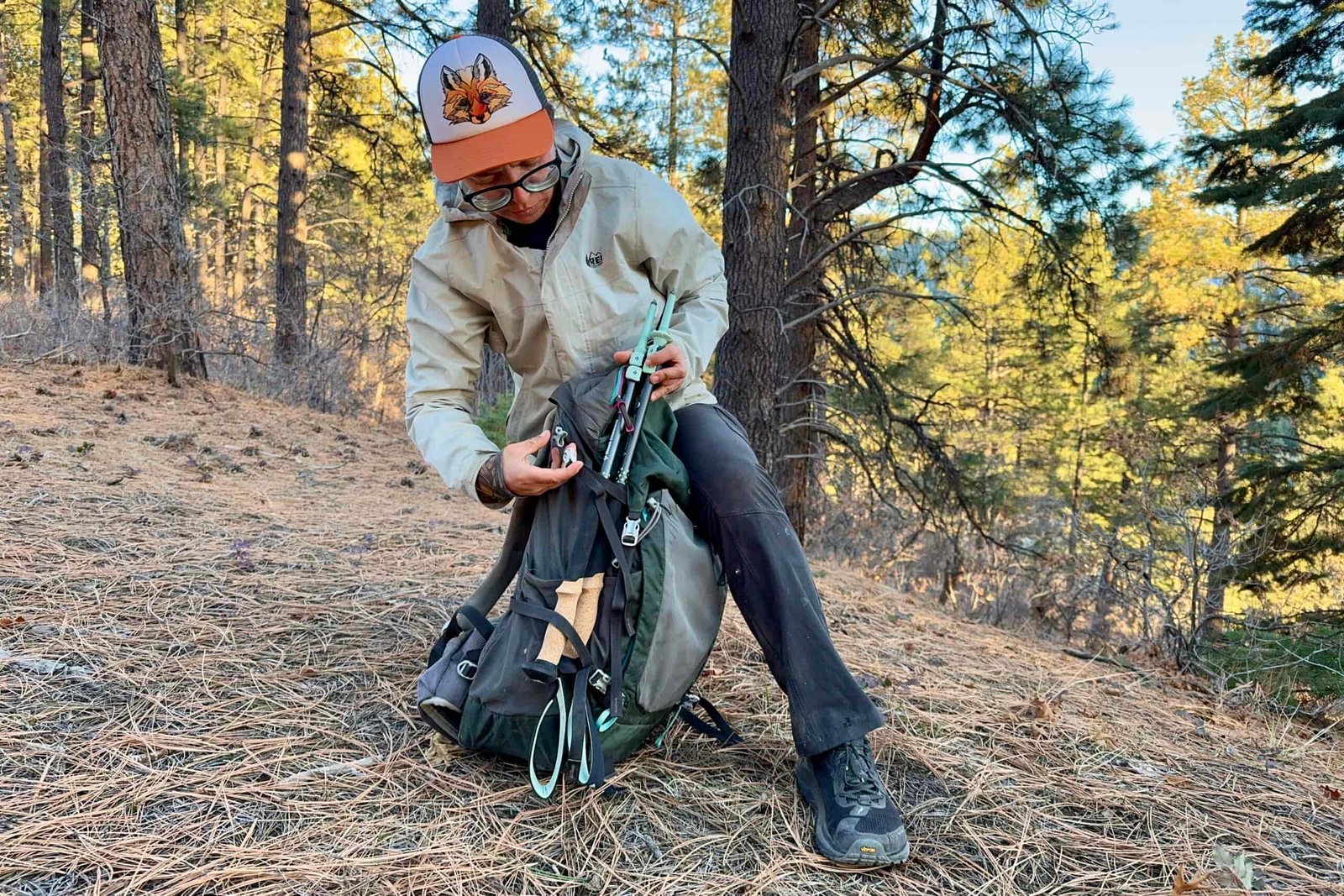 A person is clipping some trekking poles to their pack in the forest.
