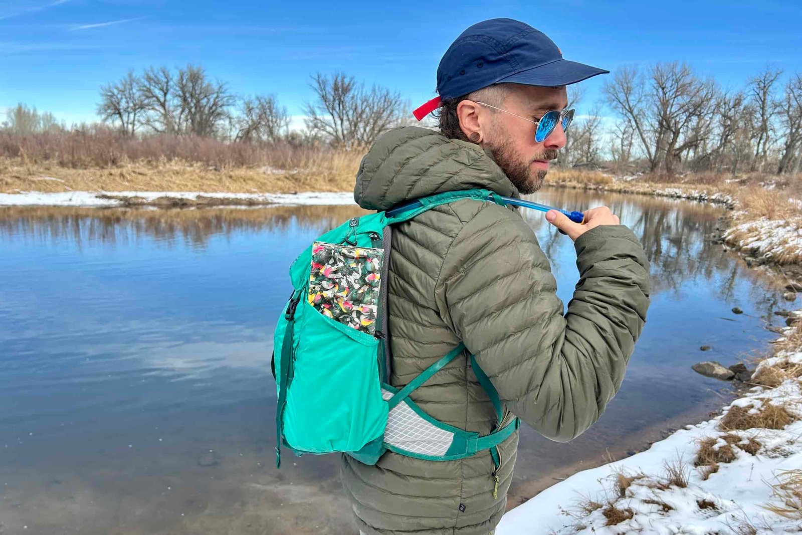 Closeup image of a man reaching for a drinking tube attached to his backpack while hiking next to a snowy river.