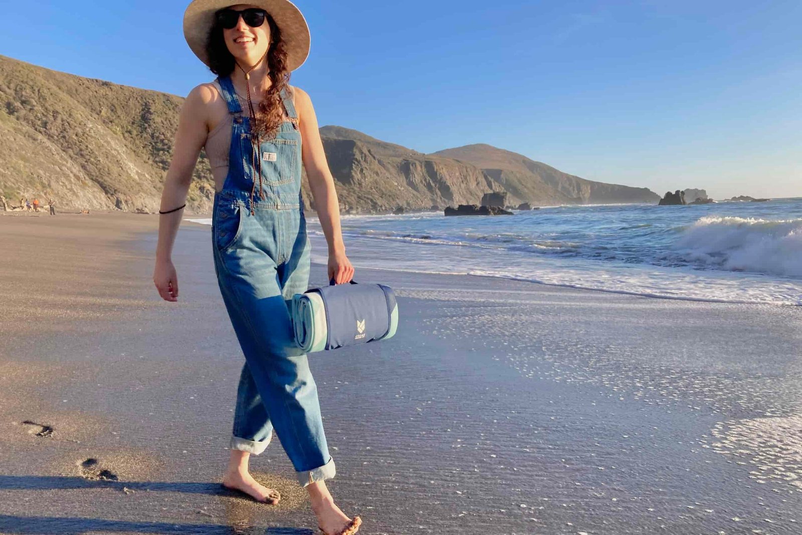 Woman walking on the beach carrying the CGear Multimats Sandlite Sand-free Mat in its handy carry case.