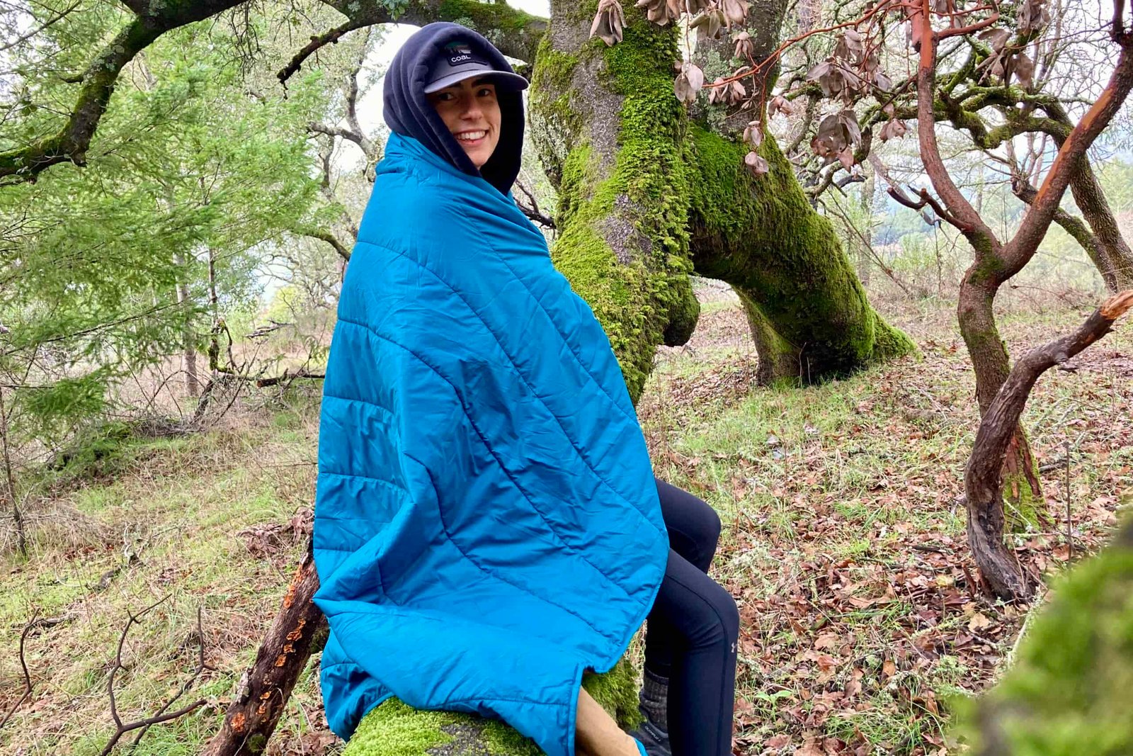 Person wrapped in the REI camp blanket while sitting on a mossy tree branch