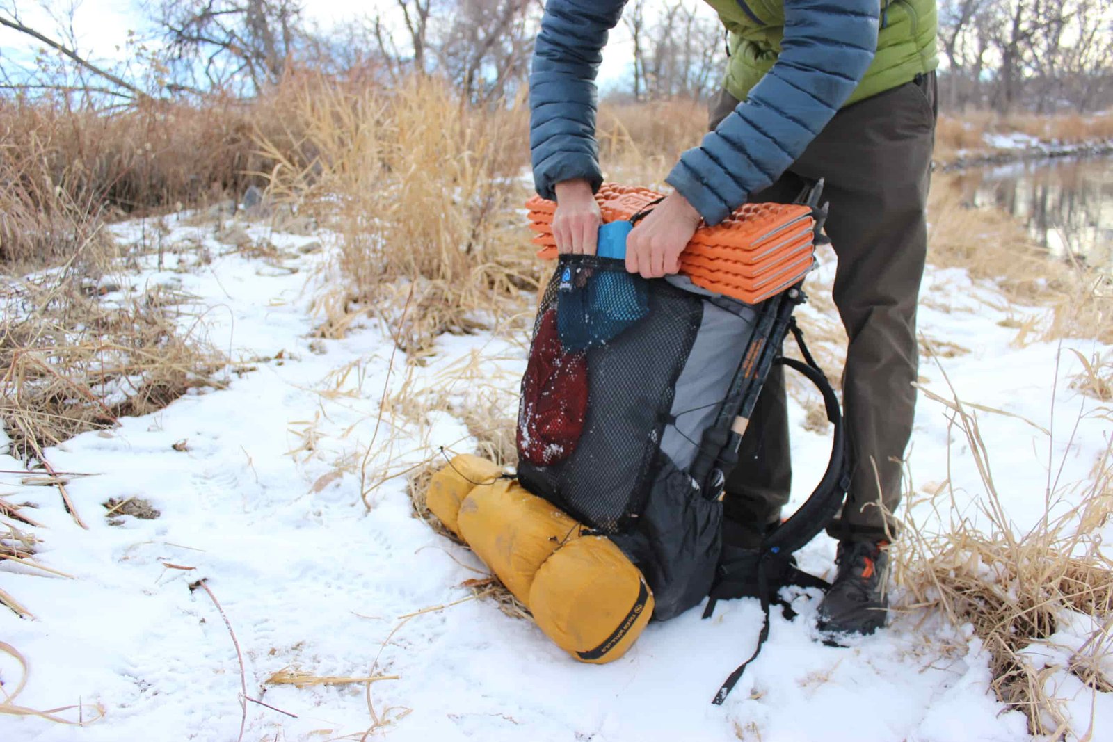 A hiker pulls a blue bag out of the front mesh pocket of a grey backpack on snowy ground.