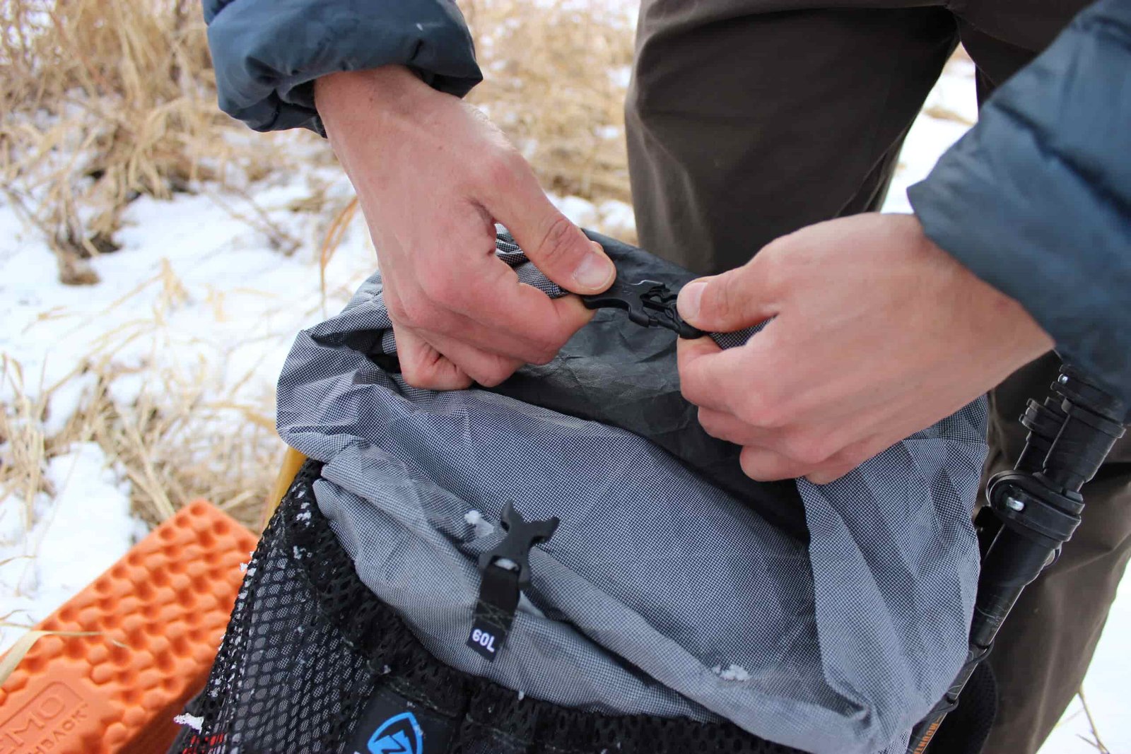 A hiker's hands buckle the roll top enclosure on a grey backpack with snow in the background.