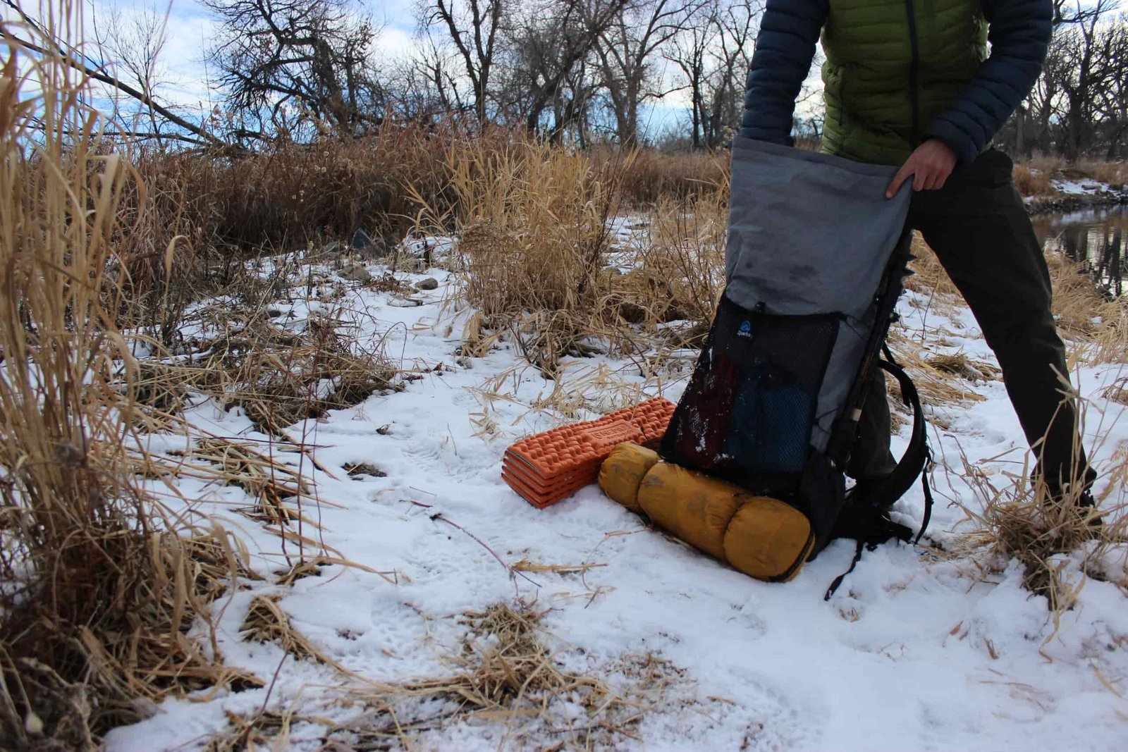 A hiker reaches into the top of a grey backpack with a tent and sleeping pad on a snowy ground.