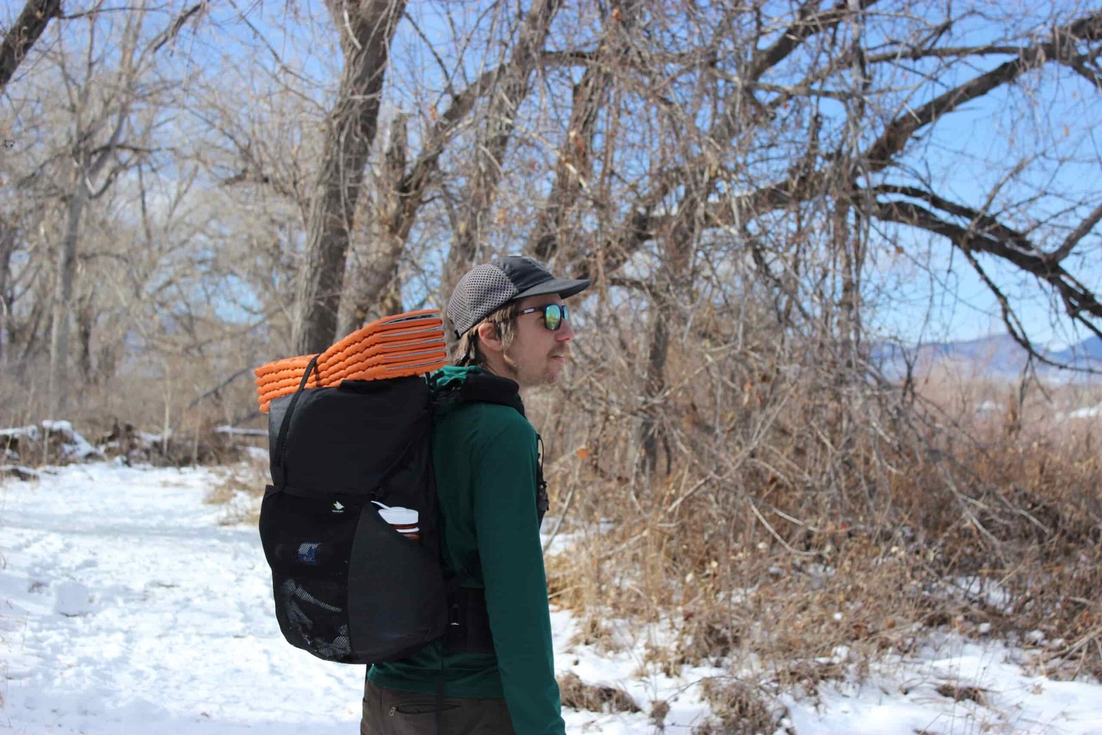 A hiker looking off into the distance while wearing the nashville pack cutaway with an orange sleeping pad on top. Trees, blue sky, and snow are in the background.