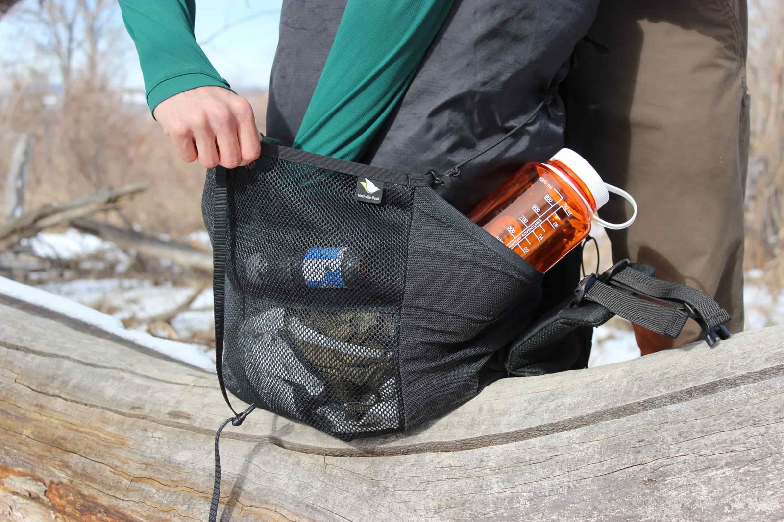 A hiker reaches into the outside pocket of a backpack with an orange water bottle in the side pocket.