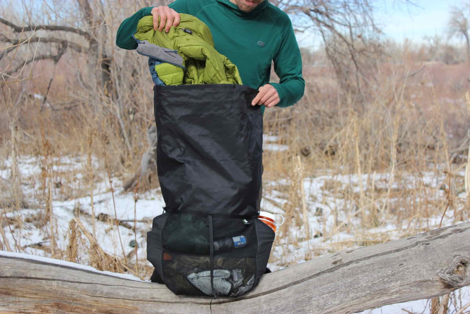 A hiker pulls a jacket out of a black backpack sitting on a log with trees and snow in the background.