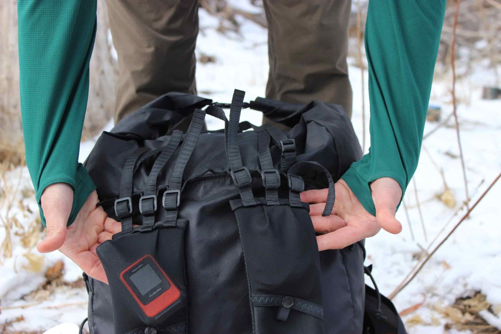 A hiker's hands displaying the shoulder strap attachments on a black backpack with a snowy scene in the background.