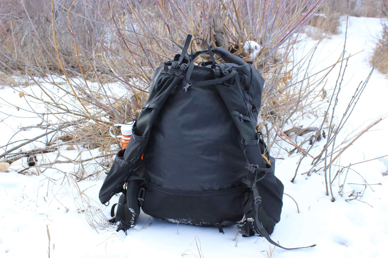 A black backpack on a snow ground with willows in the background.