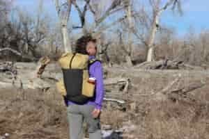 A hiker walks away from the camera wearing the pa'lante v2 backpack with trees and blue sky in the background.
