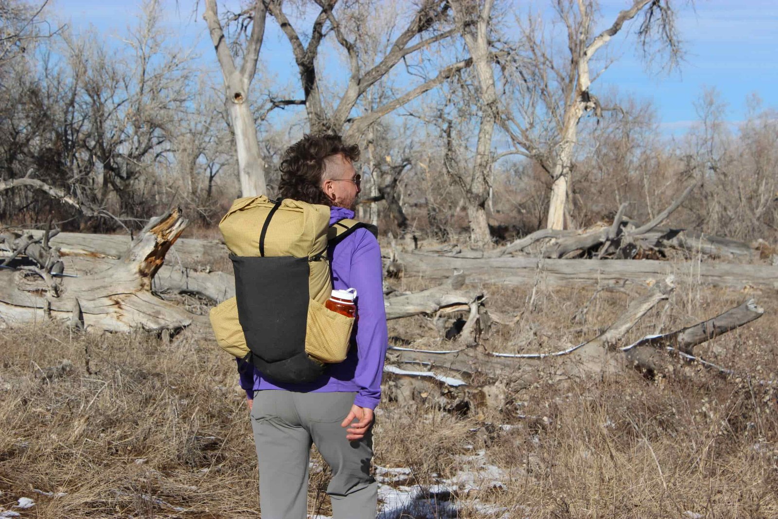 A hiker walks away from the camera wearing the pa'lante v2 backpack with trees and blue sky in the background.