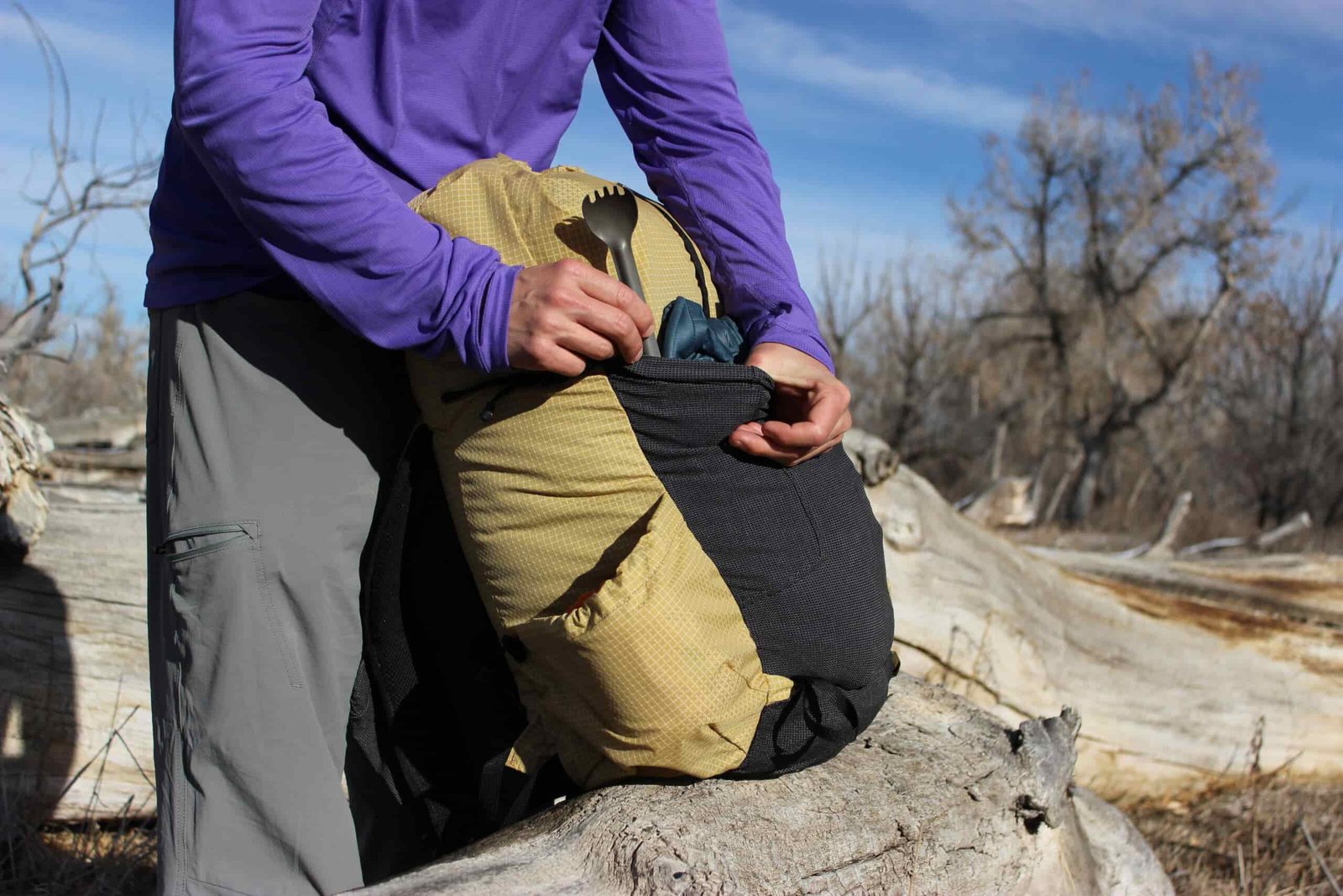 A hiker pulls a spork out of the front pocket of a yellow backpack that sits on a log.