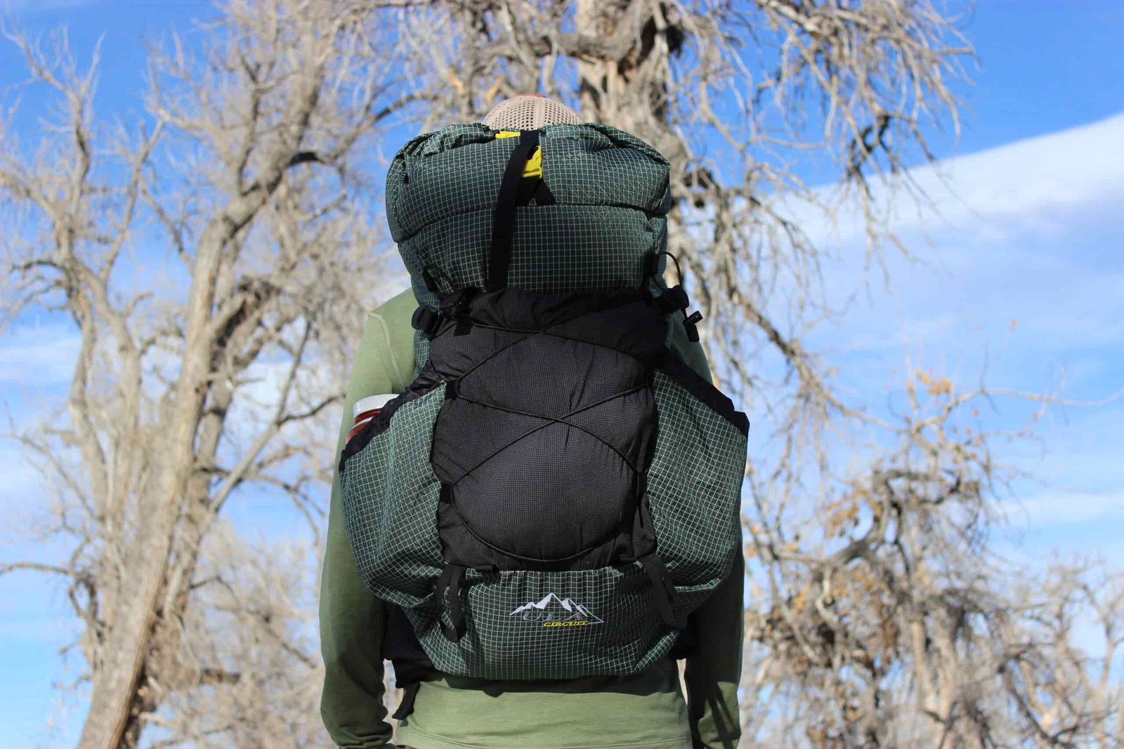 A hiker facing away from the camera while wearing the ULA Circuit backpack with trees and blue sky in the background.