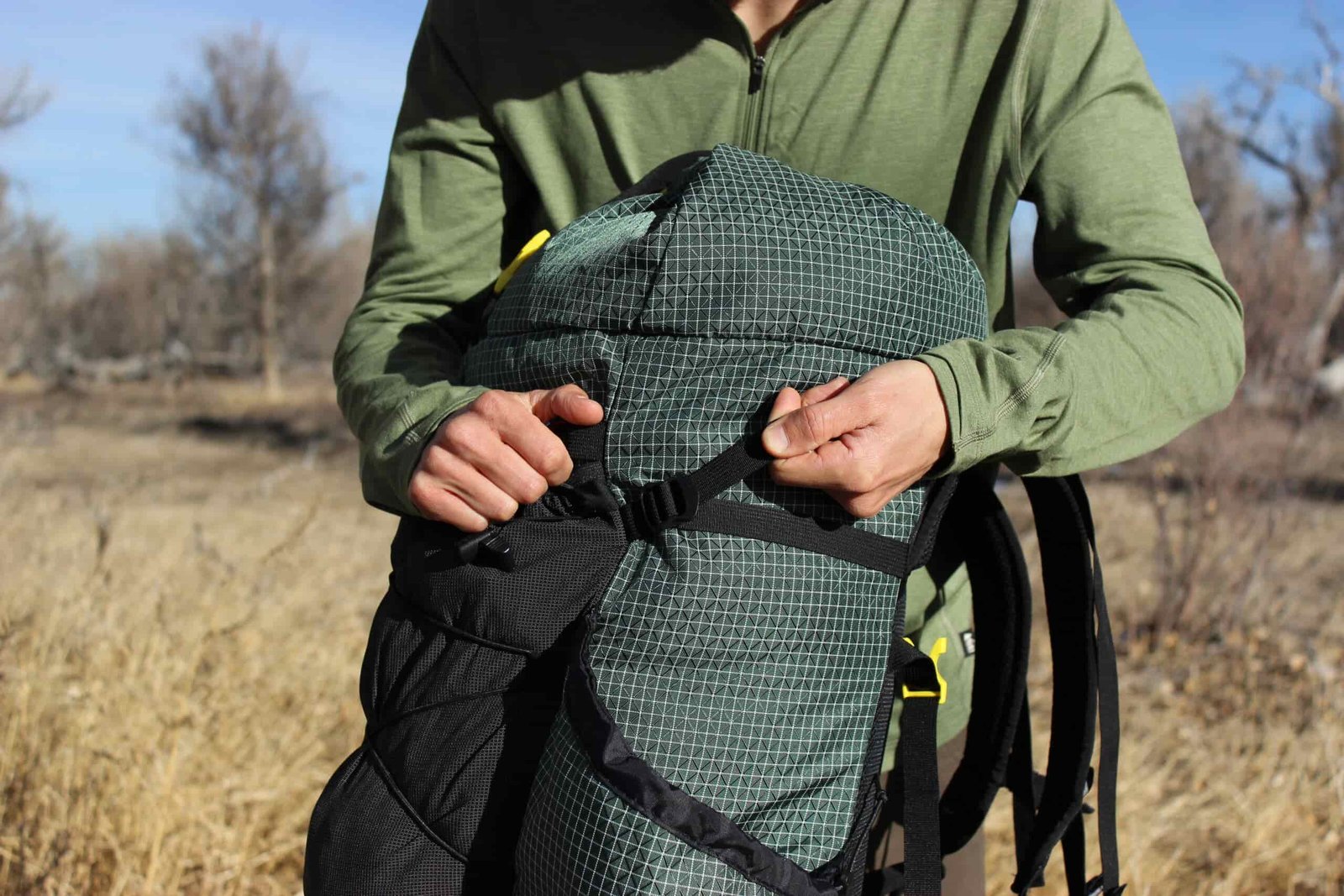 A hiker pulls on the side straps of a green backpack with trees in the background.
