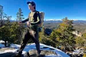 A hiker tightens the straps on a backpack while wearing it and the Smartwool Intraknit. Sky, mountains, and trees in the background.