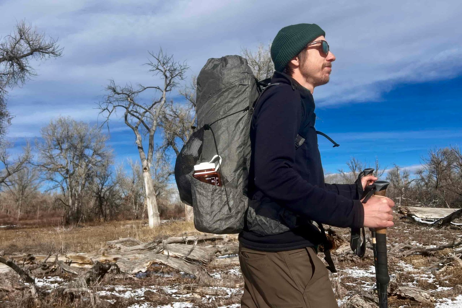 Profile view of a hiker with trekking poles carrying a grey backpacking backpack. The background is a winter scene with bare trees.