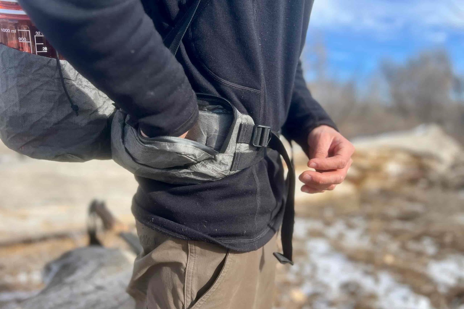 Close-up detail shot of a backpack's hip belt attachment system with a hiker's hand reaching into the hipbelt pocket.