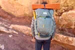 A hiker faces away from the camera while wearing a grey backpack with an orange sleeping pad on top and red rocks in the background.