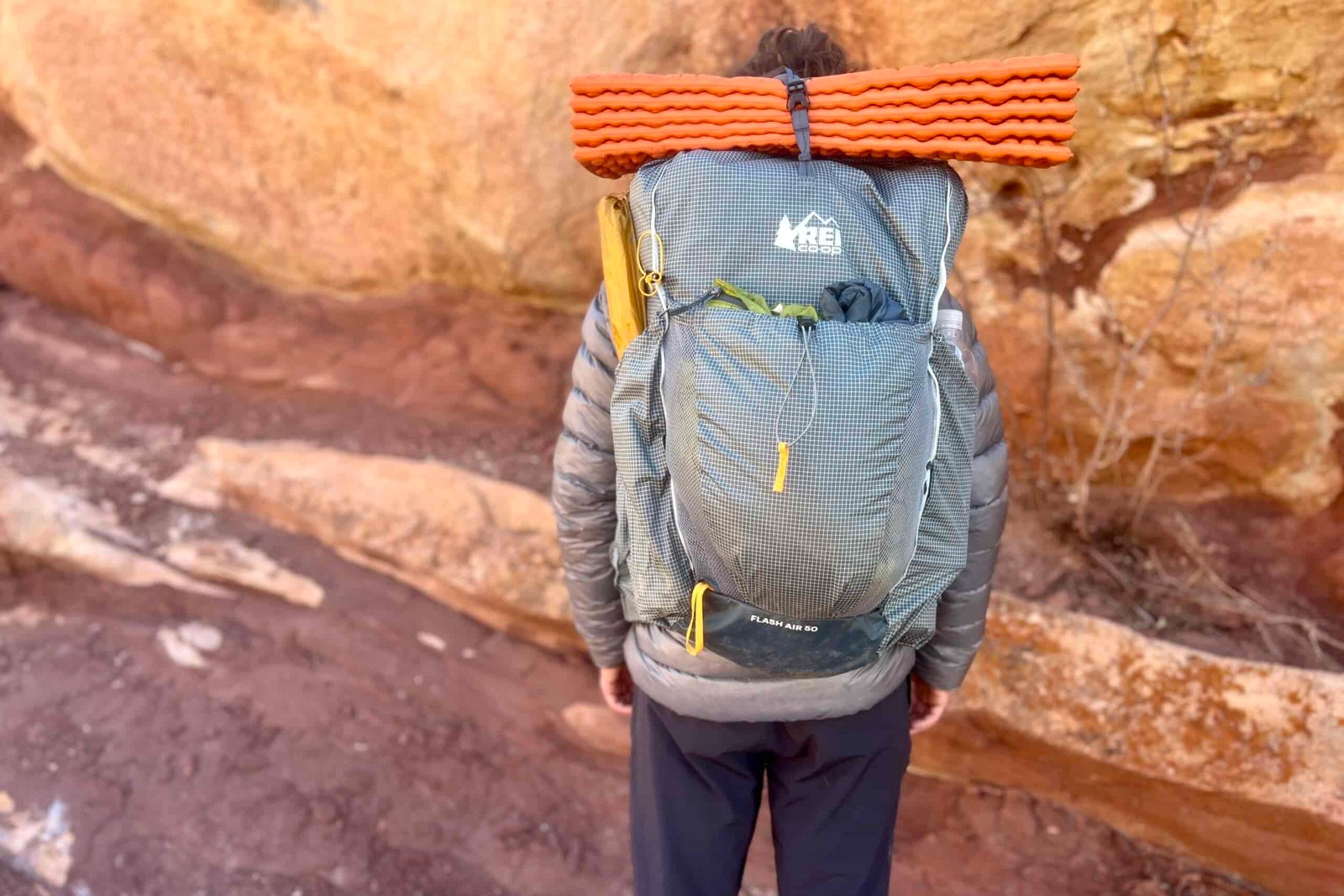 A hiker faces away from the camera while wearing a grey backpack with an orange sleeping pad on top and red rocks in the background.