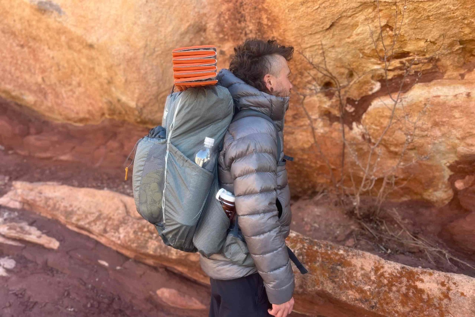 A side profile of a hiker wearing a grey backpack with an orange sleeping pad on top and red rock in the background.